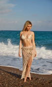 Woman in a starfish cream bikini and crochet skirt standing on a beach with ocean waves in the background