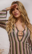 A woman wearing a long-sleeve knit dress with a vertical zigzag stripe pattern in olive green, pink, and cream, featuring a split V-neckline with a gold ring detail.