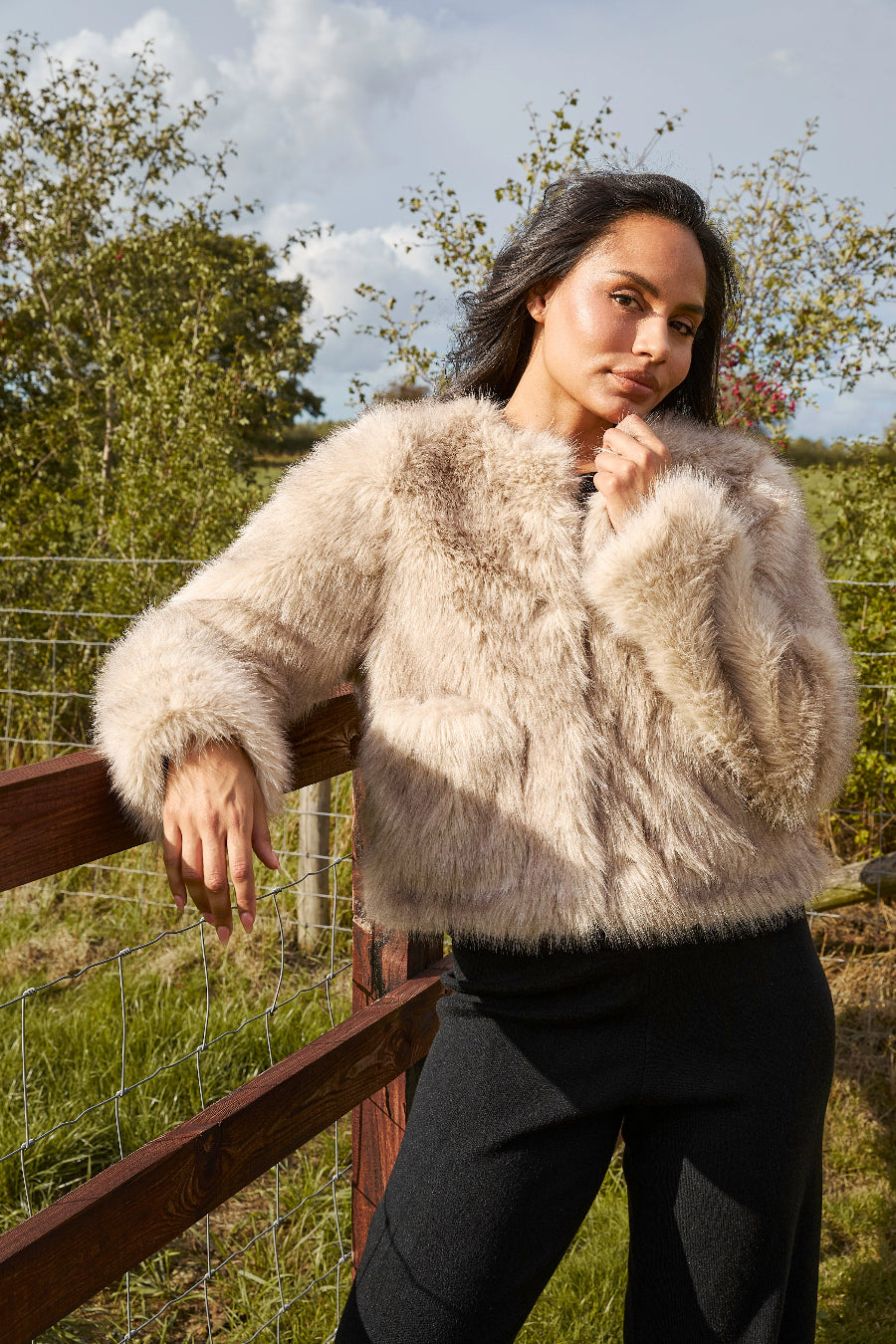 Woman wearing a fluffy beige jacket standing outdoors with trees and sky in the background